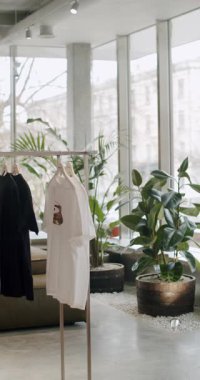 Siyah beyaz tişörtlü elbise askıları. Arka planda büyük yeşil bitkiler var. Geniş pencereli modern giyim mağazası..
