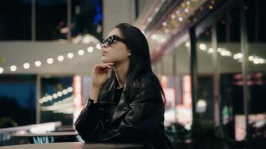 Güneş gözlüğü ve deri ceket takan şık bir kadın, arka planda bulanık şehir ışıkları ve bokeh efektleriyle iyi aydınlatılmış bir şehirde oturuyor. Gece Deri Ceketli Tarz Kadını