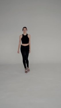 Siyah bluz ve kot giyen genç bir kadın modelin portresi, gri bir stüdyo arka planında güven içinde poz veriyor. Gri Arkaplan Karşısında Siyah Giyimli Genç Kadın. Dikey video.