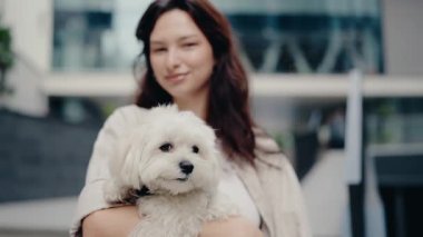 Genç bir kadın küçük beyaz köpeğini kollarında tutuyor ve kameraya gülümsüyor. Modern bir ofis binasının önünde duruyorlar. Kadın mutlu ve evcil hayvanının yanında olmaktan hoşlanıyor..