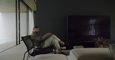 Bir adam hasır bir sandalyede geniş bir pencerenin önünde, şık, modern oturma odasındaki bir kitaba daldırılmış olarak rahatlar. Geniş Pencere 'nin kitabıyla Modern Evde Rahatlayan Adam