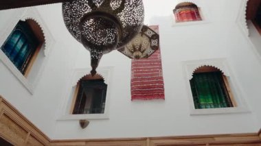 Süslü pirinç lambaları, renkli tekstil ürünleri ve kemerli pencereli beyaz duvarları olan geleneksel Fas avlusu. Bu tasarım sakin ve davetkar bir atmosfer yaratır..