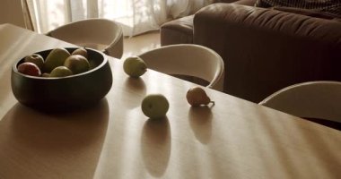 Güneş ışığı pencereden akar ve ahşap bir yemek masasını aydınlatır. Armut ve elmalarla dolu siyah bir kase süs olarak duruyor. Sıcak ışık uzun gölgeler bırakır