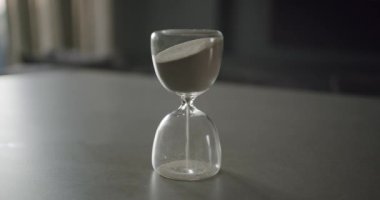 Bir el, zamanın akışını simgeleyen bir kum saatini nazikçe çevirir. İnce kum akar, kayıp giden anların görsel bir temsilini yaratır. Bu bir aciliyet hissi uyandırıyor.