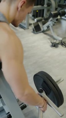 Odaklanmış genç bir adam iyi donanımlı bir spor salonundaki halterle ağırlık ayarlar, fitness bağlılığını vurgular. Modern Spor Salonu, Dikey Videoda Ağırlıkları Ayarlayan Odaklı Adam.