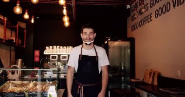 Beyaz t-shirt önlüklü, sakallı, olumlu genç erkek barista ve modern kahve evinde dururken gülümseyen ve kameraya bakan tekrar kullanılabilir yüz maskesi. Kahve dükkanında çalışırken gülümseyen genç adam..