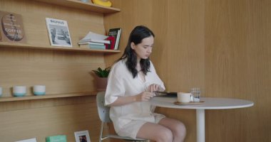 Genç bir kadın, şık dekorlar ve kitaplarla çevrili, sakin bir çalışma ortamını gösteren minimalist bir kafe masasında dizüstü bilgisayarına odaklanıyor. Kahveyi seviyor, rahat ve üretken bir sabahı somutlaştırıyor.
