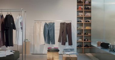 Modayı takip eden bir giyim mağazasında minimalist bir estetik sergileniyor. Ahşap raflar, metal raflar ve günlük kıyafetler. Pantolonlar, şapkalar, gömlekler ve ayakkabılar düzgünce düzenlenmiş.,