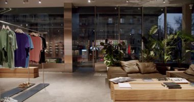 Modern kentsel sokak giyim mağazası iç mimarisi, giysi rafı, ayakkabı, rahat kanepe, ahşap masalar, dergiler, büyük pencereler ve akşam manzaralı şehir manzarası.