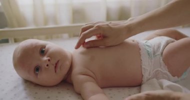 Tombul yanakları ve büyük mavi gözleri olan sevimli yeni doğmuş çocuk vücudunu hareket ettiriyor ve kameraya bakıyor. Güzel, yeni doğmuş bebek yatak çarşaflarında yatıyor, ayak parmaklarına dokunuyor gülüyor kameraya bakıyor..