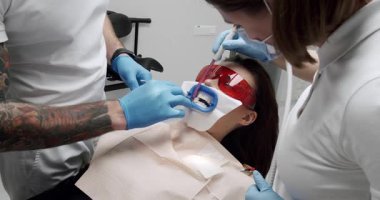 Diş hekiminin ofisinde diş beyazlatma. Modern bir klinikte ultraviyole ışık feneriyle diş beyazlatma prosedürü. Dişçi koltuğunda koruyucu gözlüklü kadın hasta..
