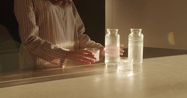 Kadın, yumuşak güneş ışığında şık bir sürahide bulunan bir bardağa tatlı su dolduruyor. Dikkatimizi bardaklara ve bardakların doldurulmasına verelim. Susuzluk, ferahlık ve sağlıklı yaşam temaları için mükemmel..