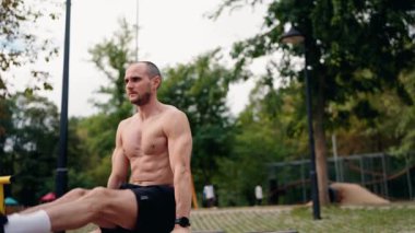 Kaslı bir adam parktaki paralel çubuklarda dibe vurur. Her tekrarı tamamlarken şekline ve gücüne odaklanıyor. Park ayarları bu fitness odaklı çekim için doğal bir zemin sağlar