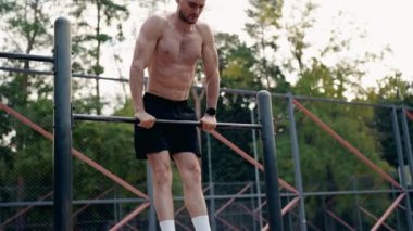Kaslı bir adam açık hava fitness parkında yatay bir çubuğa daldırıyor. Formuna ve gücüne odaklanıyor ve her tekrarı tamamlıyor. Bu da fitness yolculuğuna bağlılığını gösteriyor..