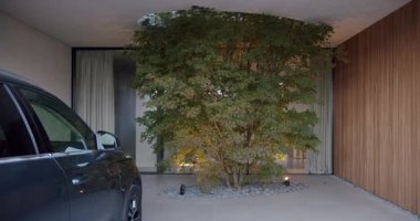 Sıcak ışıklandırma ve doğal elementlerle konforlu ve modern ev tasarımı. Garajın önüne park eden araba varış ve lüks hissi uyandırıyor..
