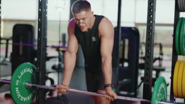 Kararlı bir erkek vücut geliştirici modern bir spor salonundaki halterlere yaslanarak ara verir. Derin nefes alıyor, bir sonraki seti için hazırlanıyor. Spor salonu ağırlıkları ve ekipmanları motive edici bir zemin sağlıyor.