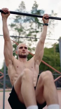 Kaslı bir adam, açık hava parkı ortamında yatay bir barda asılı duran bacağını kaldırma egzersizi yapar. Jimnastik yaparken güç ve zindelik gösteriyor..