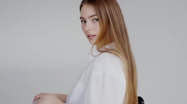 Gür beyaz bluzlu, uzun sarı saçlı genç bir kadın yumuşak gri arka planda zarafet ve tefekkür sergiliyor. Beyaz Bluz Giyen Zarif Genç Kadın Basit Arkaplan Üzerinde