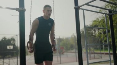 Sisli bir sabahta açık hava fitness parkında jimnastik halkaları kullanan odaklanmış genç bir sporcu. Eğitimde kararlılık ve güç. Spor salonunda jimnastik yüzükleri eğitimi alan genç adam.