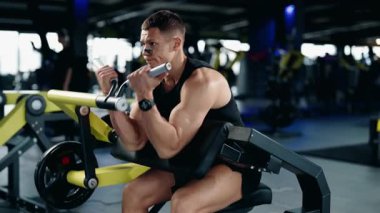 Kaslı bir adam modern bir spor salonundaki bir makinede biseps yapıyor. Formuna ve gücüne odaklanıyor, fitness 'e olan bağlılığını gösteriyor. Spor salonu temiz ve iyi donanımlı.,