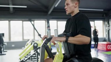 Odaklanmış genç bir adam, modern bir fitness merkezinde azim ve güç gösteren bir spor makinesinde çalışıyor. Bir Spor Makinesine Yoğun Odaklanmış Genç Adam Eğitimi