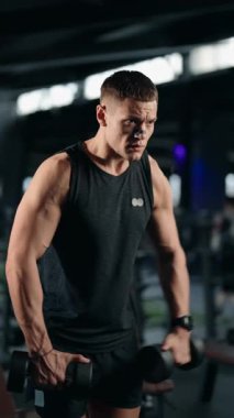 Kas yığını bir adam modern bir spor salonunda dambıllarla yanal yükseltme yapar. Egzersizine odaklanmış ve fiziğini geliştirmeye kararlı. Spor salonu iyi donanımlı ve koyu renkli.,