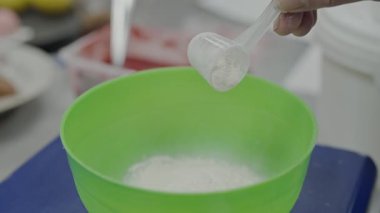Bir insan yeşil bir karıştırma kabına ustalıkla şeker döker, muhtemelen lezzetli bir tatlı için malzemeleri hazırlar. Kurulum, tatlı yapımına odaklanmış bir mutfak ortamı öneriyor..