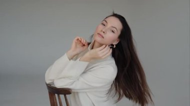 Elleri boynunda, sakince sandalyede oturan bir kadının videosu. Stüdyoda beyaz kazaklı ve pantolonlu güzel esmer kadın poz veriyor..