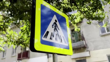 Şehirdeki yaya geçidi tabelası. Sarı çerçeveli mavi-beyaz yol tabelası. Gökyüzü ve ağaçlar işarete yansıyor. Yol güvenliği ve trafik yönetmelikleri kavramı. Şehir sokak sahnesi.