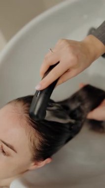 Bir kadın saçını kuaförde yıkatıyor. Stilist saçlarını ılık suyla yıkarken nazikçe kafa derisine masaj yapar. Yakın çekim, lüks ve rahatlatıcı deneyimi vurgular..