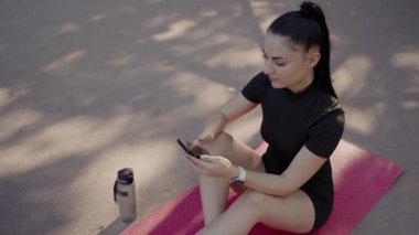 Odaklanmış genç bir kadın parkta pembe bir yoga minderine oturur ve spor seansından sonra akıllı telefonunu kullanır. Yanında su şişesi vardı. Genç Kadın Yoga minderinde otururken Smartwatch 'ini kontrol ediyor