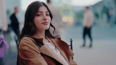Kahverengi ceketli, uzun saçlı genç bir kadının portresi, arka planda bulanık ışıklarla şehir ortamında dışarıda oturuyor. Genç Kadın Dışarda Poz Veriyor