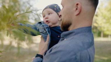 Genç, sakallı bir baba oğlunu sevgi dolu bir şekilde parkta tutuyor ve hassas bir aile anı yaşıyor. Genç Baba bebeğini parkta kucağında tutuyor.