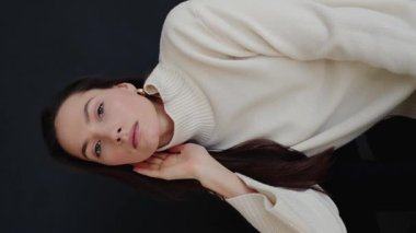 Stüdyoda poz veren beyaz kazaklı ve pantolonlu güzel esmer kadın. Güzel bir kadının pürüzsüz ve sağlıklı bir teni var. Cilt bakımı, yüz gerdirme, kozmetik. Dikey video.