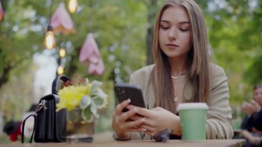 Bir kafede akıllı telefonuna odaklanmış genç bir kadın, canlı bir şehir parkı ortamı ile çevrili. Harmanlama teknolojisi, eğlence, kentsel yaşam tarzı. Bir kafede akıllı telefon kullanan genç bir kadın.