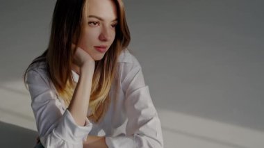 Beyaz bluzlu, düşünceli genç bir kadın, sakin bir ifadeyle içeride oturuyor, yumuşak, doğal ışıkla işaretlenmiş. Beyaz tişörtlü düşünceli genç kadın Hafif Işıkta oturuyor