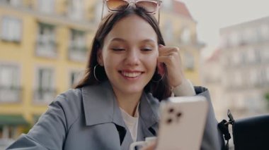 Güneş gözlüğü takan neşeli genç bir kadın canlı kentsel binaların arka planına karşı telefonuyla video görüşmesi yapıyor. Genç Kadın Renkli Şehir Ayarlarında Video Görüşmesinden Hoşlanıyor