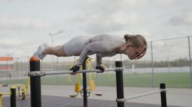 Açık hava spor salonunda paralel barlar üzerinde zor bir egzersiz yapan odaklanmış genç bir kadın esneklik ve güç gösterisi yapıyor. Atletik Kadın Açık Spor Salonu 'nda Gelişmiş Antrenman Yapıyor