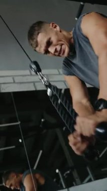 Odaklanmış bir adam, modern bir spor salonundaki bir kablo makinesinde yoğun bir ağırlık çalışması yapıyor. Kararlı ifadesi ve kaslı fiziği fitness ve eğitime olan bağlılığını yansıtıyor. Dikey