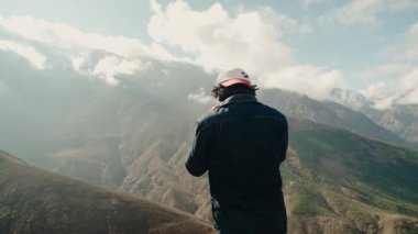 Beyzbol şapkası ve kot ceket giyen bir adam Fas 'ın görkemli dağ manzarasını seyrediyor. Önünde engebeli bir manzara uzanır bulutlar tepelerin üzerinde sürüklenir..