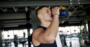 Formda genç bir adam, iyi donanımlı bir spor salonunda çalışırken su şişesiyle su içer. Odaklanma ve sağlığını gösterir. Modern Spor Salonunda Çalışırken Su İçen Genç Sporcu