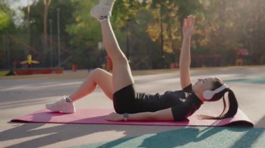 Spor kıyafetlerine odaklanmış genç bir kadın açık havada esneme egzersizleri yapıyor. Parkta kulaklıklarla güneşli bir günün tadını çıkarıyor. Parkta güneşli bir günde dışarıda egzersiz yapan genç bir kadın.