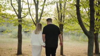 Genç bir çift, sonbahar ağaçları ve yaprakların arasından süzülen yumuşak güneş ışığı ile çevrili sessiz bir parkta el ele dolaşır. Bir yakınlık ve bağlantı anı yakalıyorum..