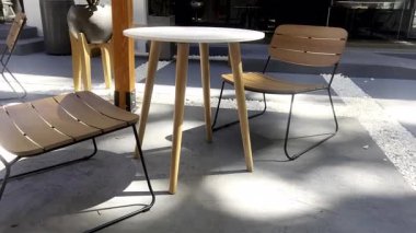 Açık hava kafe mobilyaları ahşap sandalyeler ve beyaz bir masa içerir, ahşap bir pergola altında gölgeler bırakır. Modern sandalyeler ve masa müşteriler için bir karşılama alanı oluşturur..