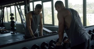 Spor salonunda dambıllarla çalışan odaklanmış genç erkekler modern fitness ortamında kendini adamışlık ve güç gösterisi yapıyorlar. Modern Spor Salonu 'nda Ağırlık Kaldıran İki Adam Odaklanıyor