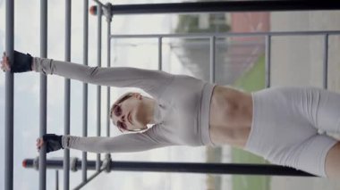 Genç, atletik bir kadın spor yapmadan önce, şık ve modern bir spor kıyafeti giyer. Şehir ortamını gören büyük pencerelerin önünde duruyor ve sağlık sorunlarını vurguluyor.