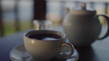 Ön planda dumanı tüten bir kahve fincanı ve arka planda hafifçe bulanık bir çaydanlık içeren rahatlatıcı bir görüntü. Sakin bir sabah ya da sakin bir kahve molası verir..