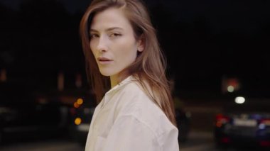 Beyaz gömlekli şık bir genç kadın, gece şehir arkaplanıyla poz veriyor, dinginlik ve güven taşıyor. Beyaz Gömlekli Genç Kadın Gece Düşünceli Poz Veriyor