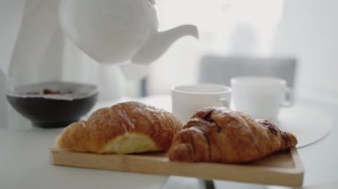 Sabah kahvaltısı taze demlenmiş çayla birlikte fincana dolduruluyor, yanında da masada kruvasanlar var. Sıcak, doğal bir ışıkla güne başlamak huzurlu bir atmosfer yaratır..