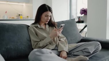 Genç bir kadın, modern bir oturma odasındaki bir kanepede rahat bir şekilde dinlenirken görülüyor, akıllı telefonunu kullanmaya kendini kaptırmış. Atmosfer sakin ve sade..